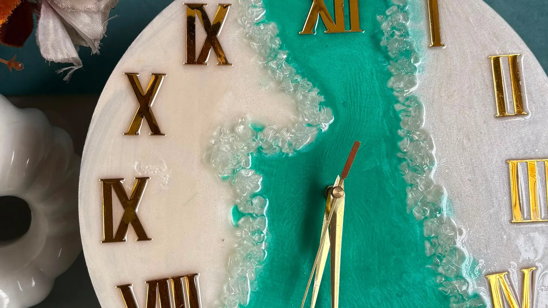 Macro detail of the ocean-pour clock face: turquoise resin meeting pearl white, with a ridge of crushed glass forming the foam line between them and gold Roman numerals set into the surface