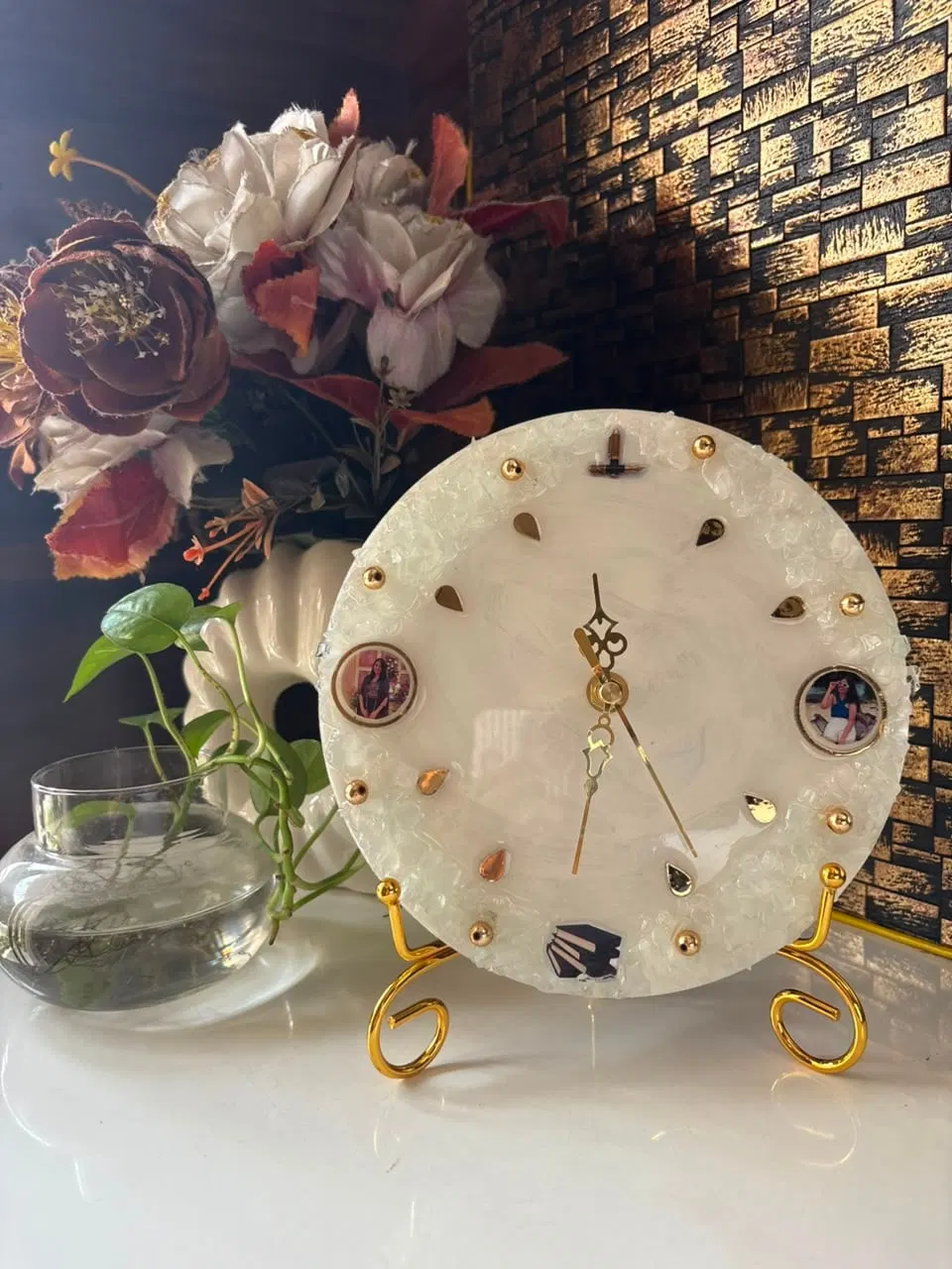 A pearl-white resin clock with crushed glass at the rim, gold markers and two small round photographs set into the face, resting on a gold stand beside dried flowers
