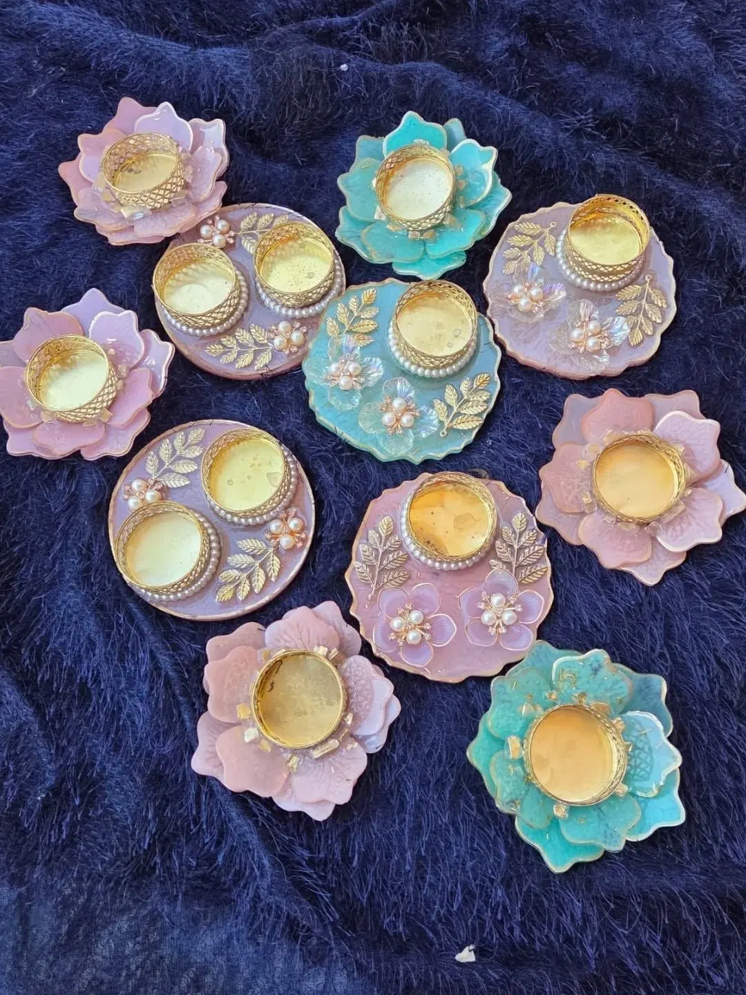 Eleven resin tealight holders and pooja thalis laid out on dark blue fur: flower-shaped ones in blush pink and mint, round thalis with gold leaf sprigs and pearl-and-crystal flowers, each holding a filigree brass tealight cup
