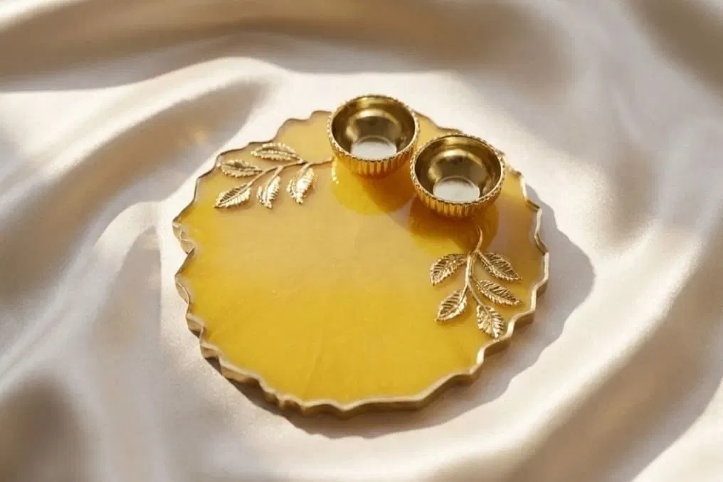 A round marigold-yellow resin pooja thali with a scalloped gilded edge and gold leaf sprigs set into the surface, holding two small fluted brass diyas, photographed on cream silk