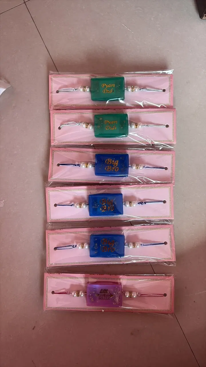 Six packaged resin rakhis laid out in a row: two green Pyari Didi, three blue Big Bro and one lilac Little Sister, each strung with pearls on a blush card