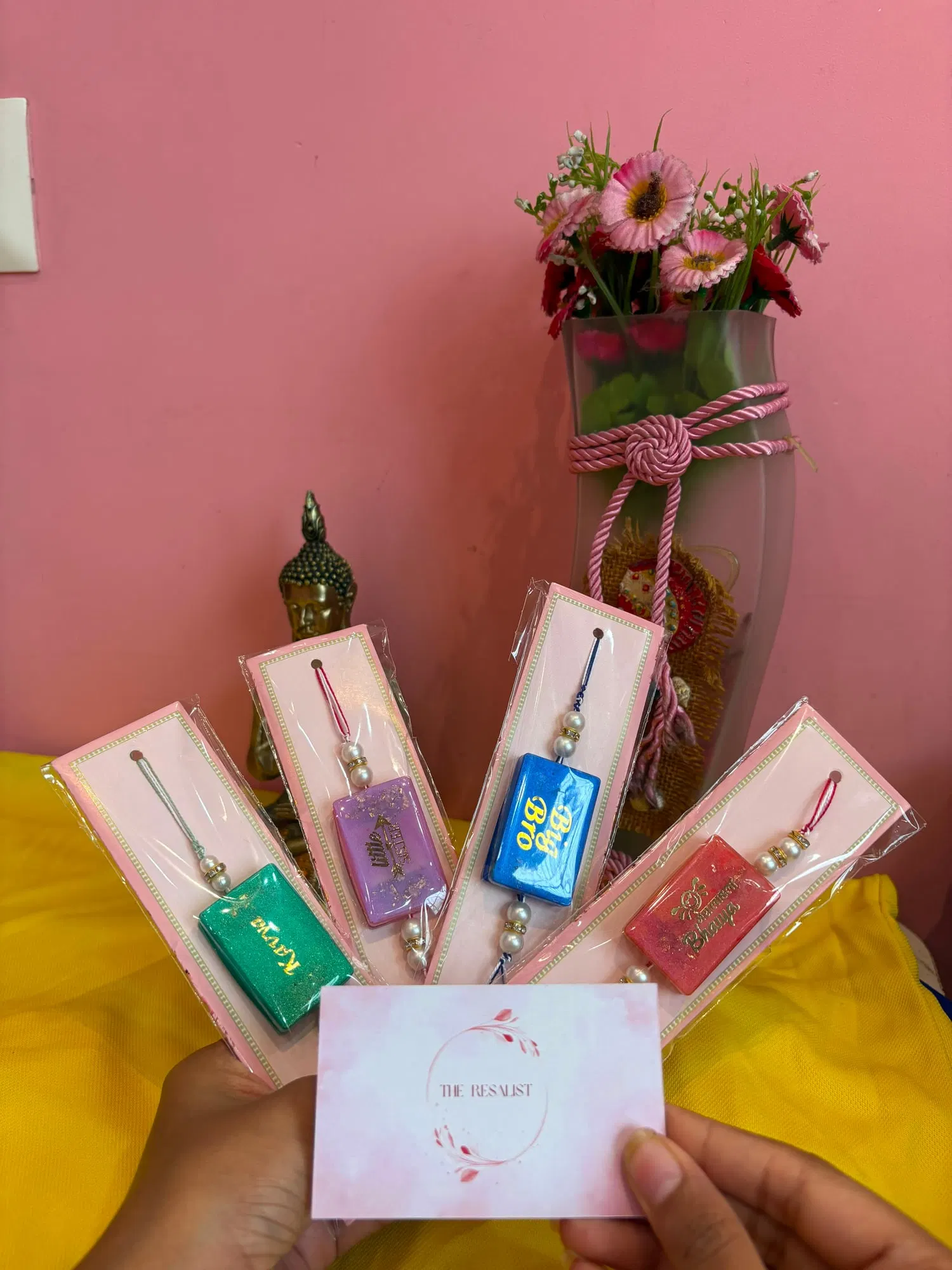 Four packaged resin rakhis in green, lilac, blue and coral with gold lettering, fanned out above The Resalist brand card