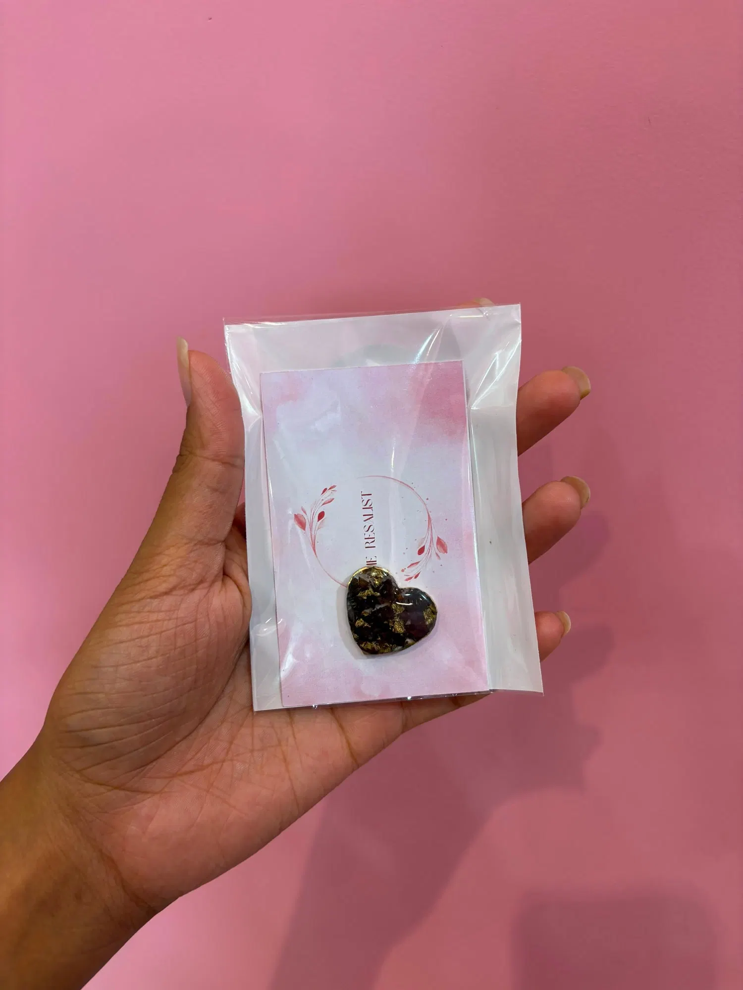 A small heart pendant of preserved dark rose petals and gold leaf in resin, sealed in a clear sleeve over The Resalist brand card