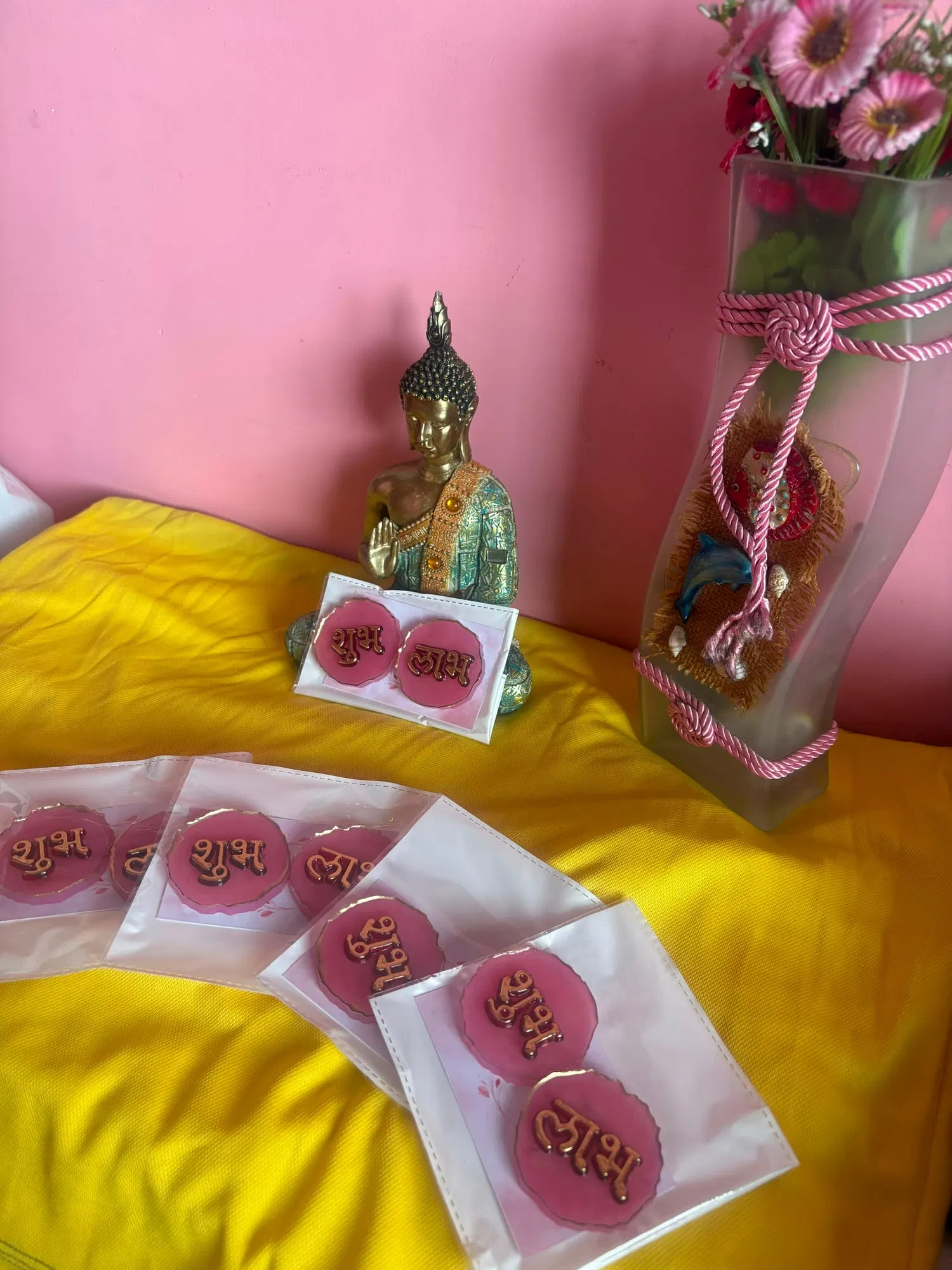 Packaged pairs of rose-pink resin discs with gilded edges, each embossed with the Hindi words Shubh and Labh in gold, arranged on yellow cloth beside a brass Buddha and pink flowers