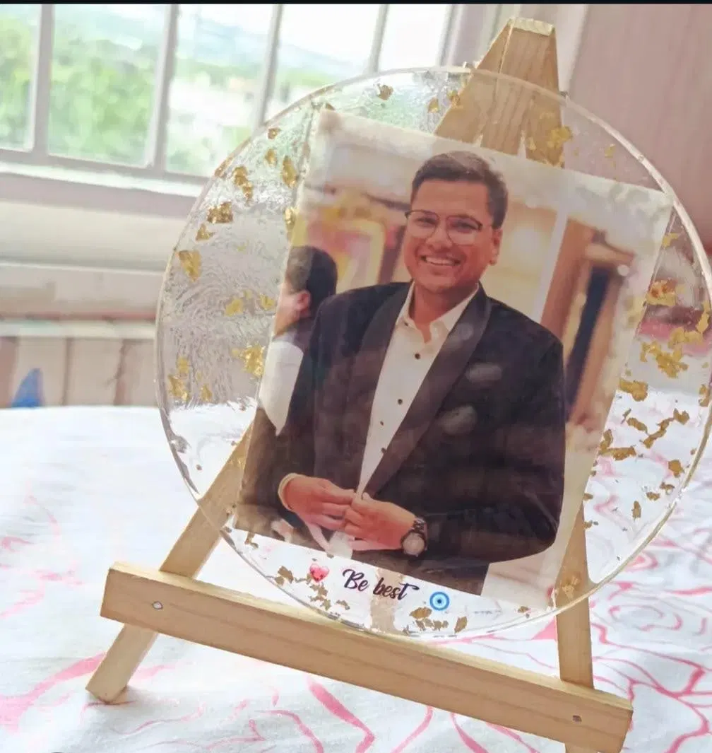 A transparent round resin frame with gold foil flakes suspended in it, holding a portrait photograph, standing on a wooden easel