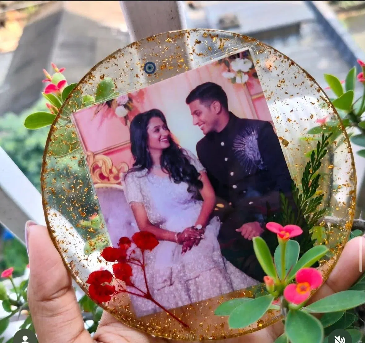 A round gold-flecked resin frame holding an engagement photograph, with real dried red flowers and greenery set into the resin at its edge