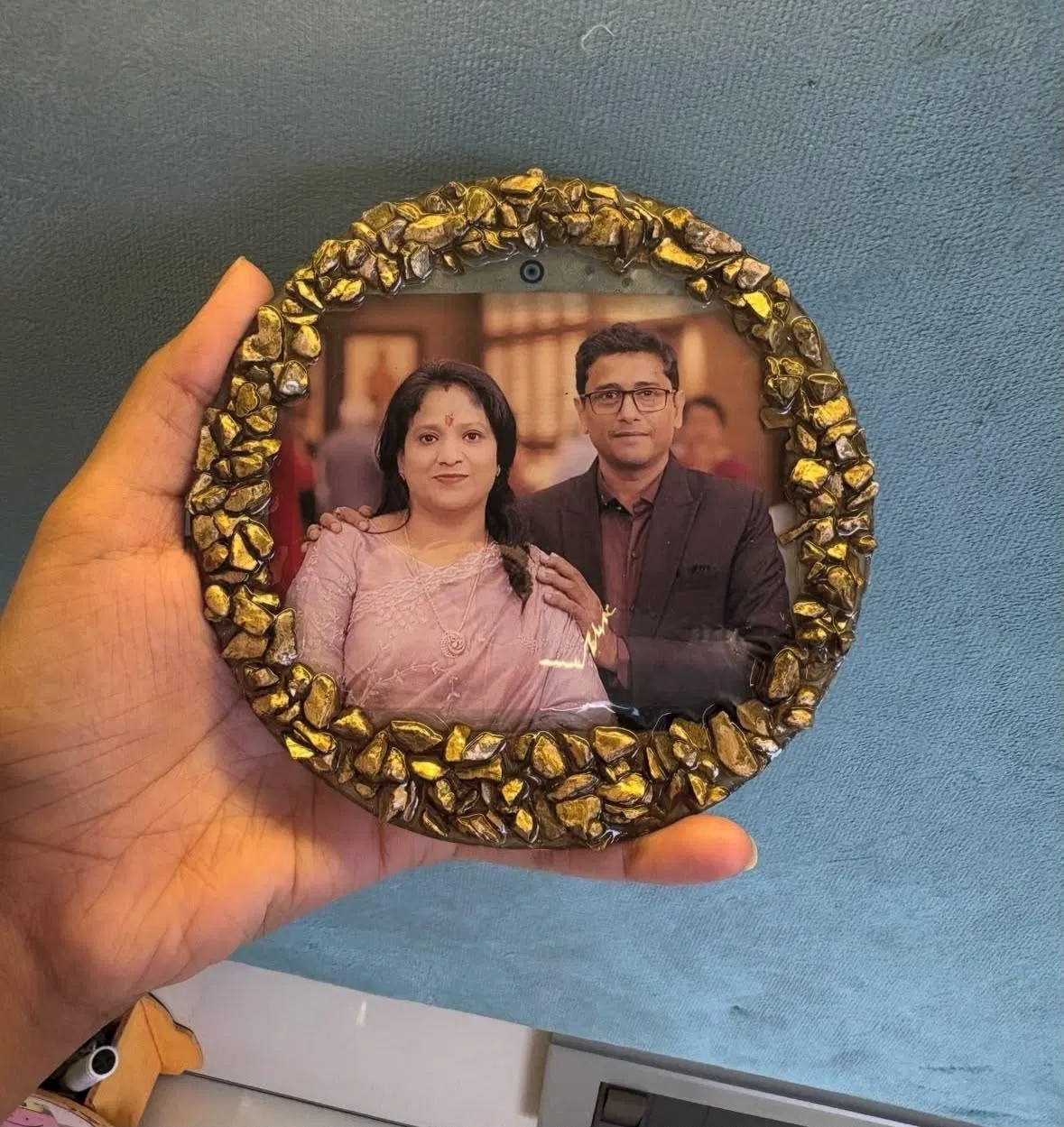 A round resin frame ringed with gilded stone chips, holding a photograph of a couple, held in a hand