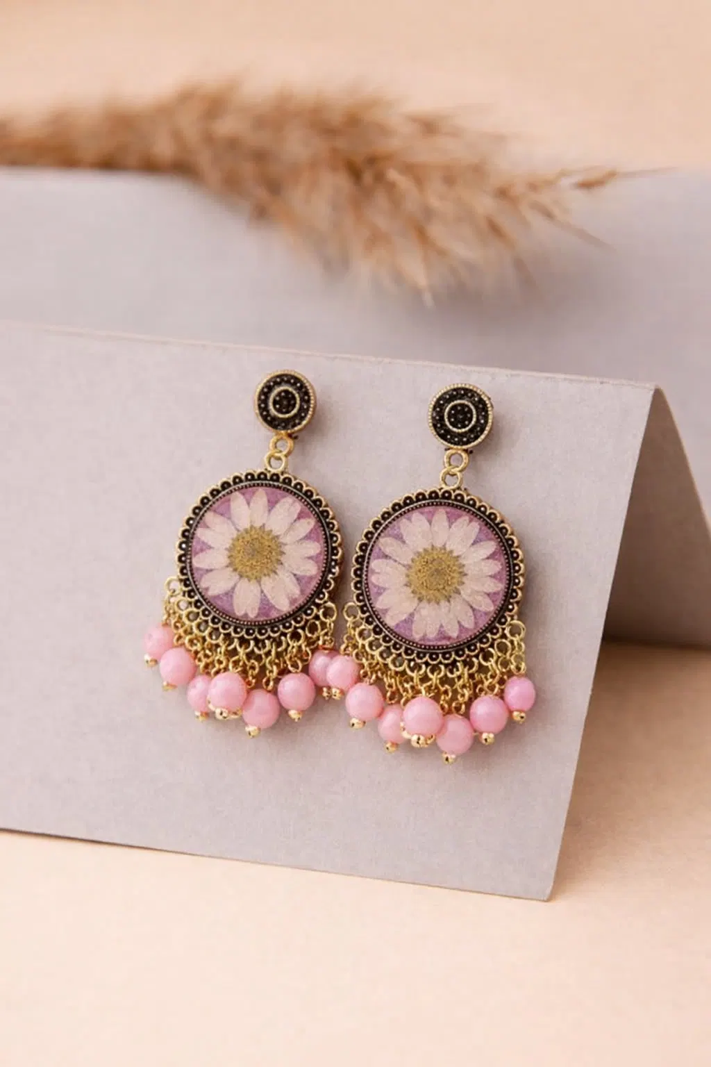 Antique gold jhumkas with a single pressed white daisy on a lilac resin base, finished with a row of pink bead drops, propped on kraft card