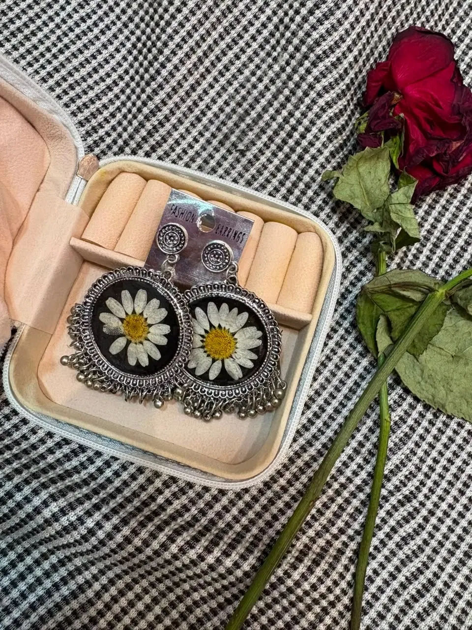Silver jhumkas with white pressed daisies set against deep black resin, sitting in a blush jewellery case beside a dried red rose