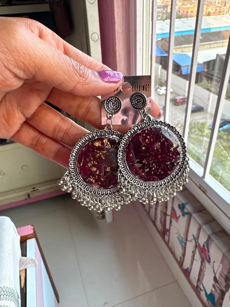 Round silver jhumkas filled with deep maroon crushed rose petals and gold flecks in resin, held near a window