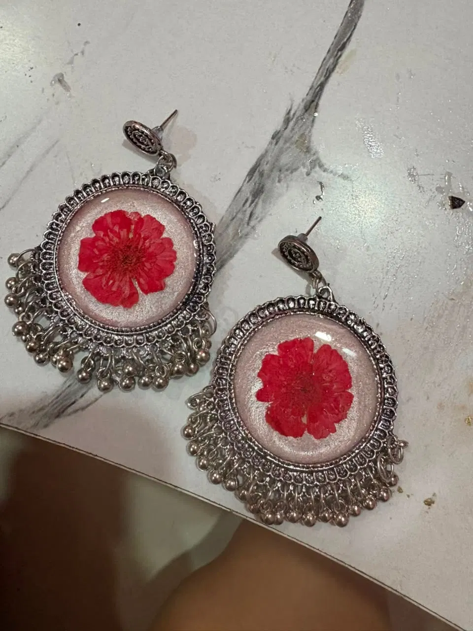 Silver jhumkas each holding one bright red pressed bloom on a pearlescent resin base, on a marble surface