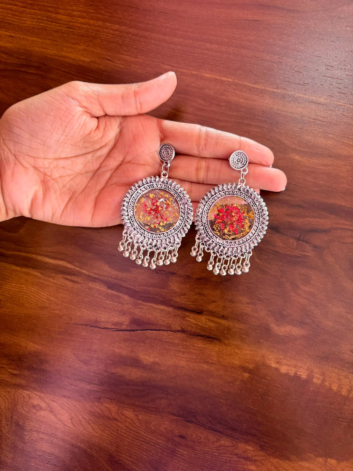 Round oxidised silver jhumkas holding a red dried flower and gold leaf suspended in clear resin, held above a dark walnut table