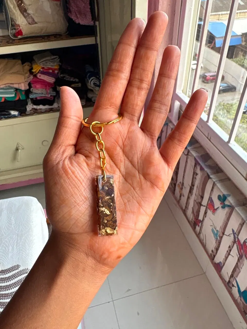 A slim rectangular resin bar keychain filled with dried rose and gold flakes, on a gold chain in an open hand