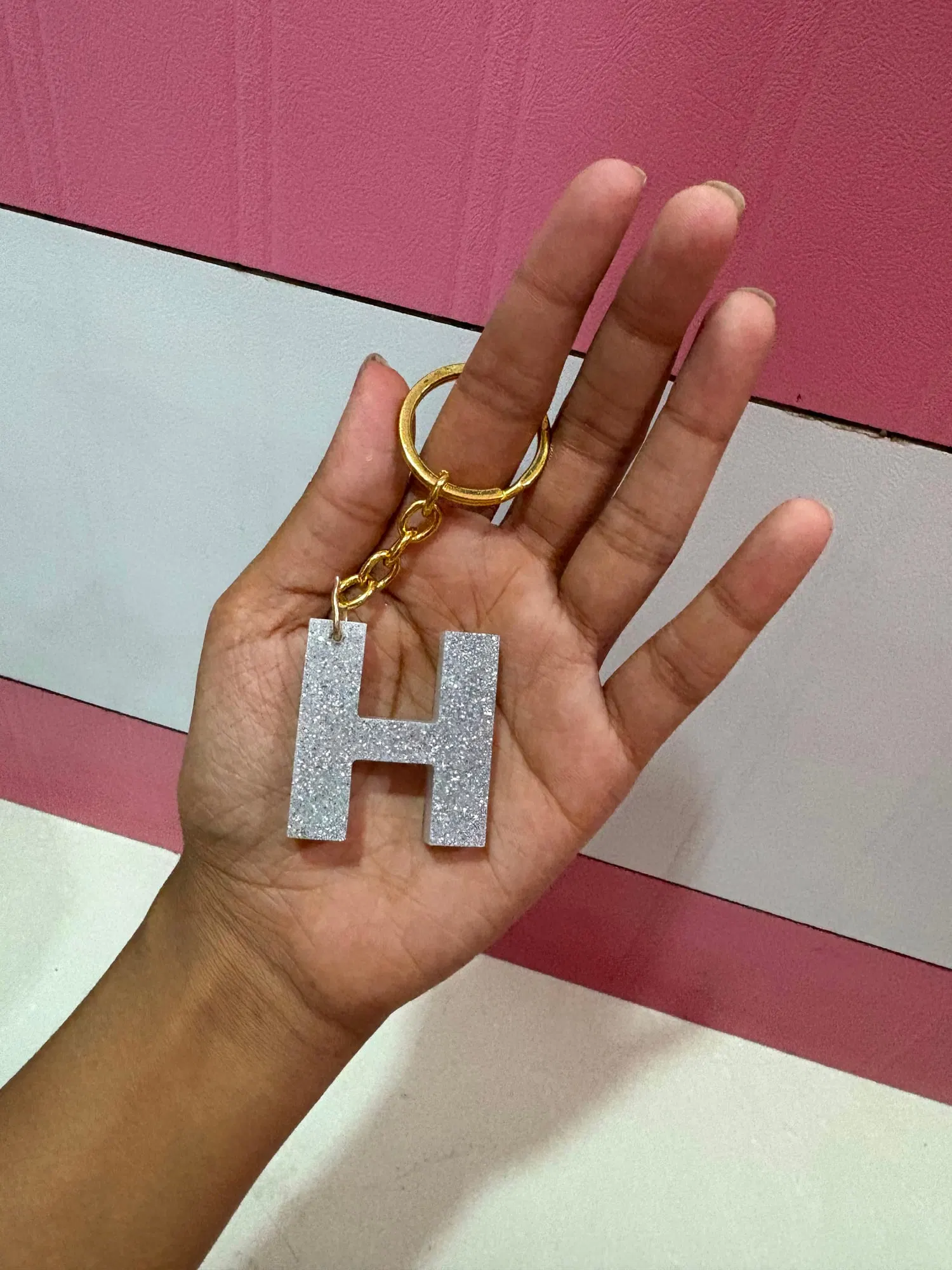 A chunky letter H keychain in white and silver glitter resin on a gold ring, held in an open palm