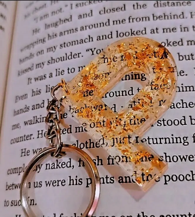 A clear resin letter R keychain filled with gold leaf flakes, lying on the open page of a book