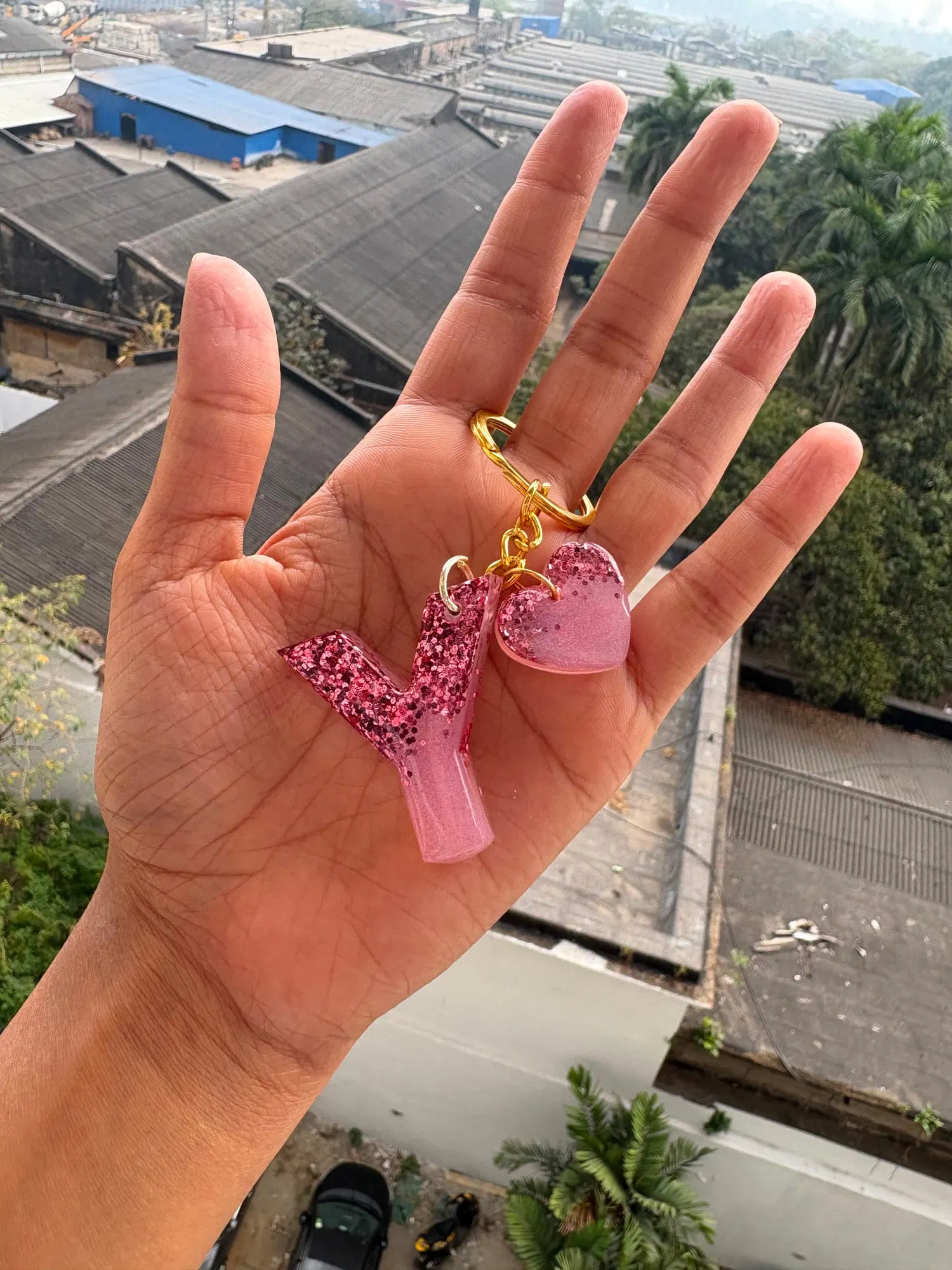 A pink glitter letter Y keychain with a matching pink heart charm, held up in a palm above a rooftop view