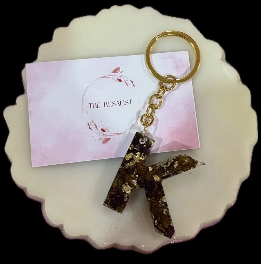 A resin letter K keychain made from real preserved rose petals and gold leaf, on a gold chain, resting on a white resin coaster beside The Resalist brand card