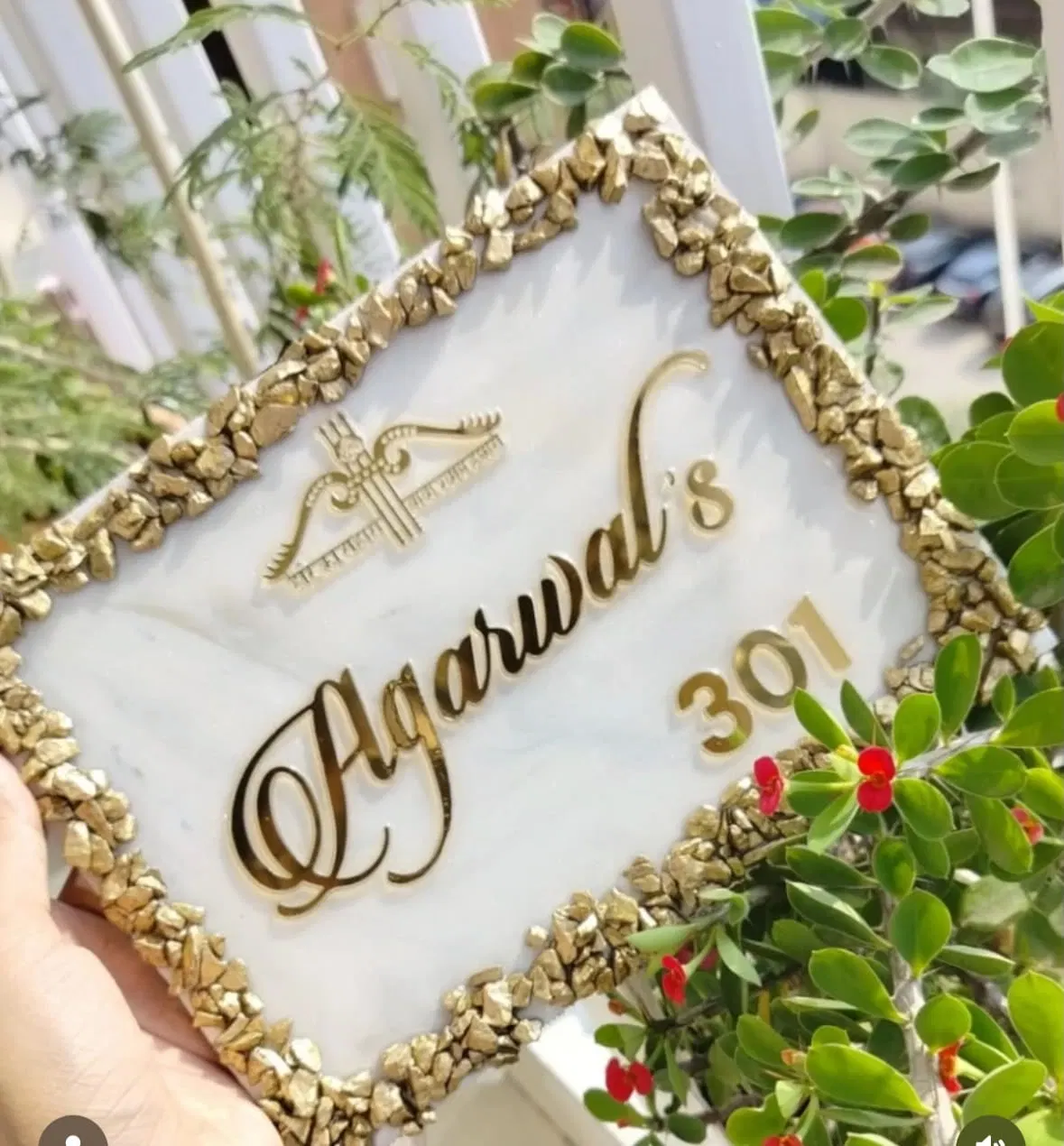 A white marble resin name plate reading Agarwal's 301 in gold script with a gold religious emblem, framed by a complete border of gilded stone chips, held up outdoors among green leaves