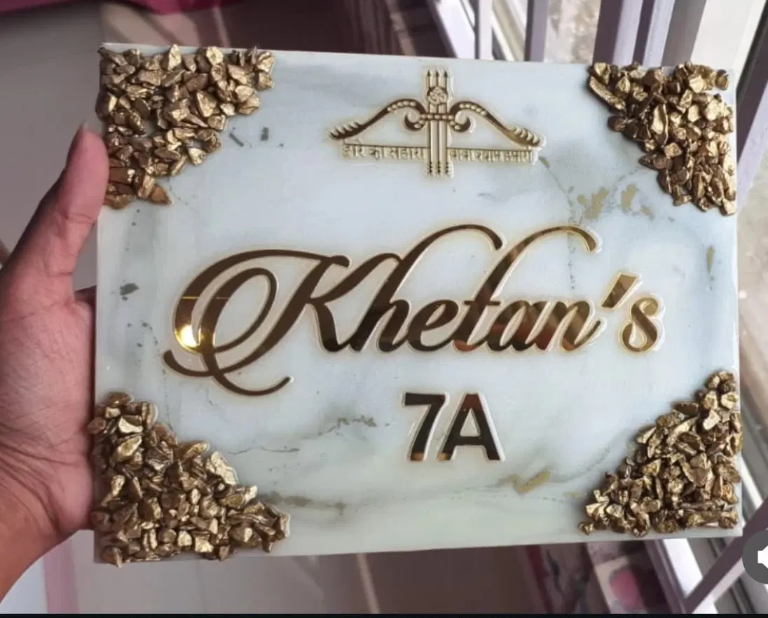 A pale marble resin name plate reading Khetan's 7A in gold script, topped with a gold religious emblem and edged with gilded stones