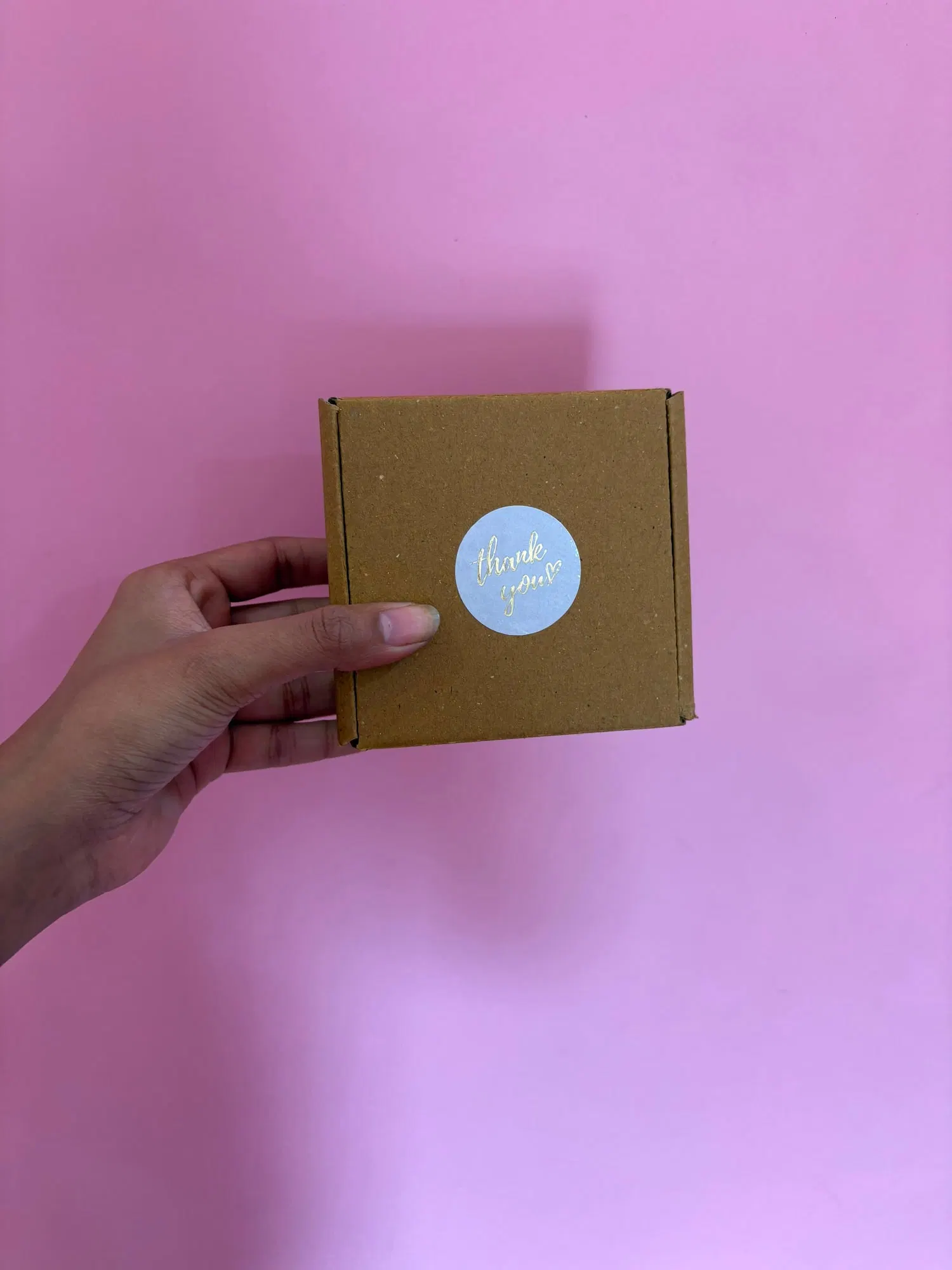 A small kraft box sealed with a gold-foil thank-you sticker, held against a pink wall