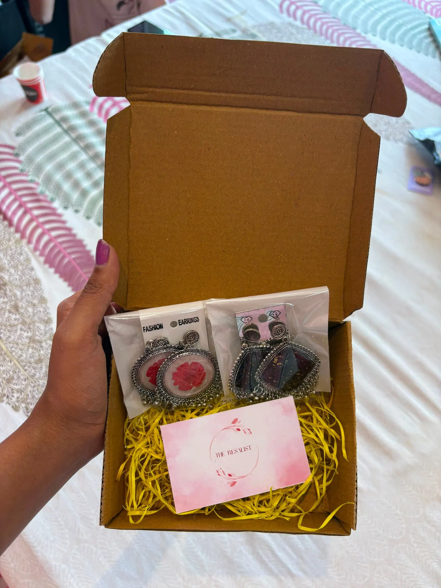 An open kraft box with two pairs of resin jhumkas in sealed sleeves resting on yellow shredded paper above the brand card