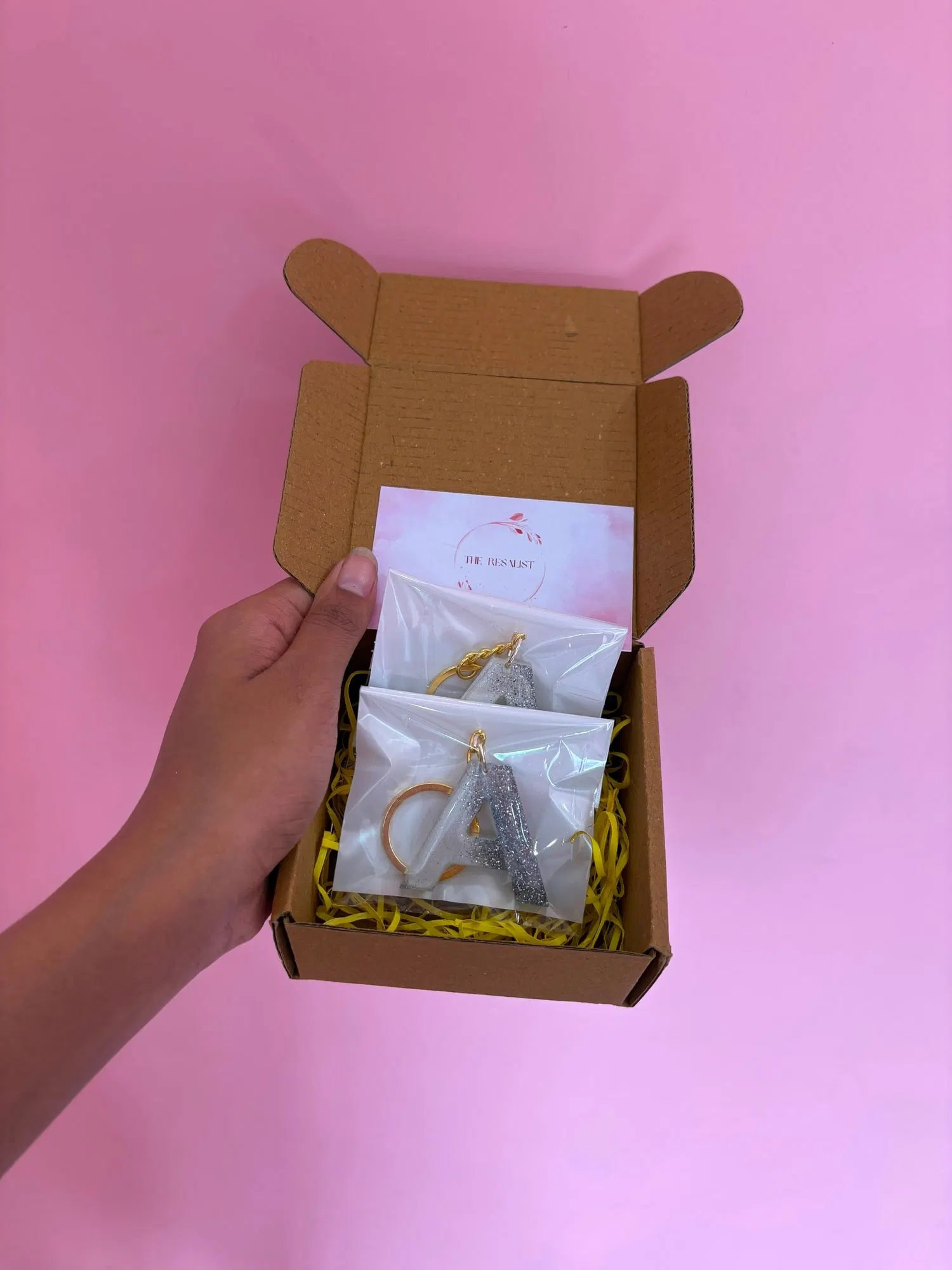 An open kraft box lined with yellow shredded paper, holding two letter keychains in frosted sleeves and The Resalist's blush brand card