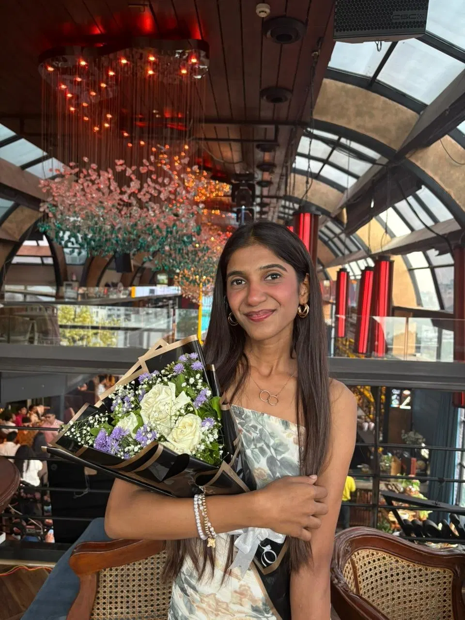 One of the sisters holding a bouquet of white roses and purple blooms in a glass-roofed cafe