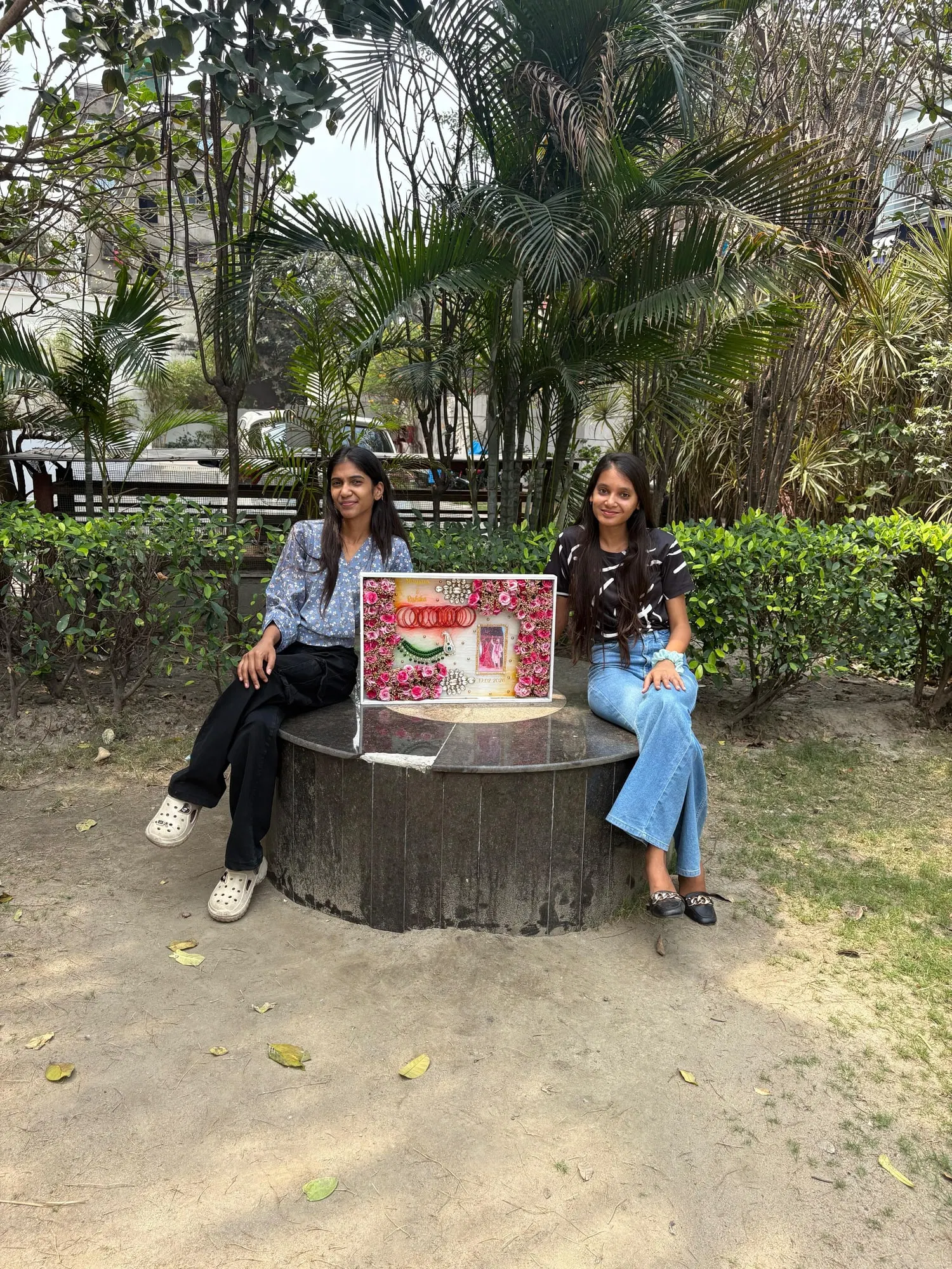 Two sisters sitting on either side of a large finished varmala preservation frame, outdoors in a garden with palm trees behind them