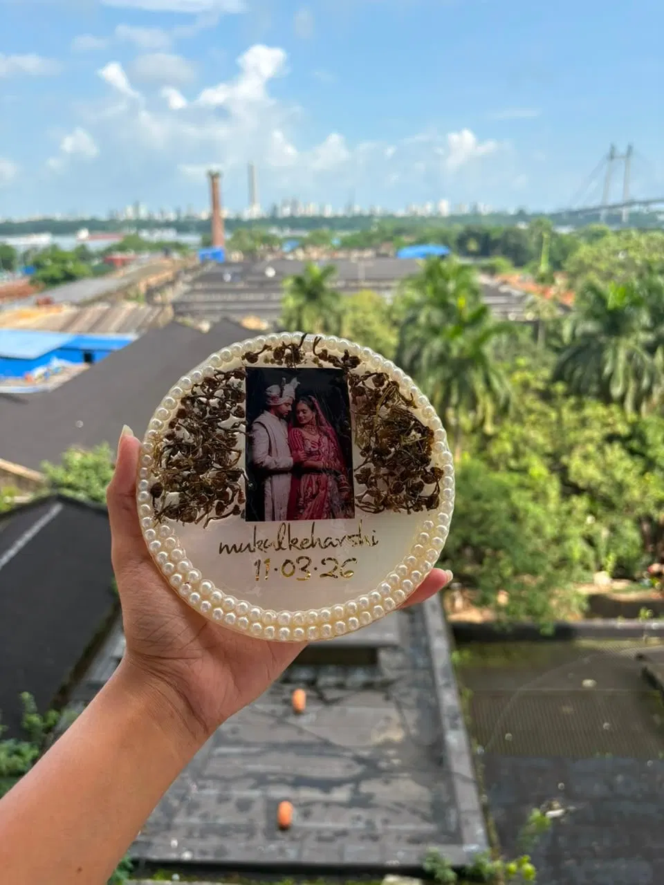 A six-inch round resin keepsake edged with pearls, holding dried varmala buds around a couple's wedding photo with their names and date in gold script, held up against the Kolkata skyline