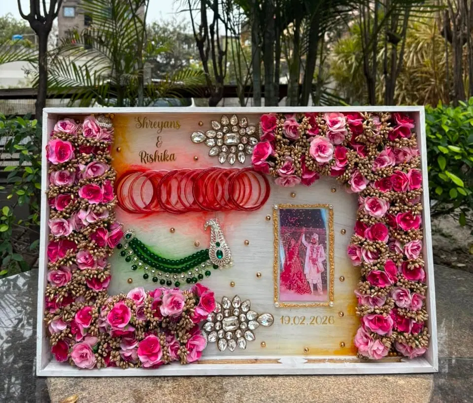 A large white shadow-box frame holding a full preserved pink-and-gold varmala around the couple's wedding photograph, with red bangles, a green bead necklace, kundan brooches, their names and the date 19.02.2026 in gold