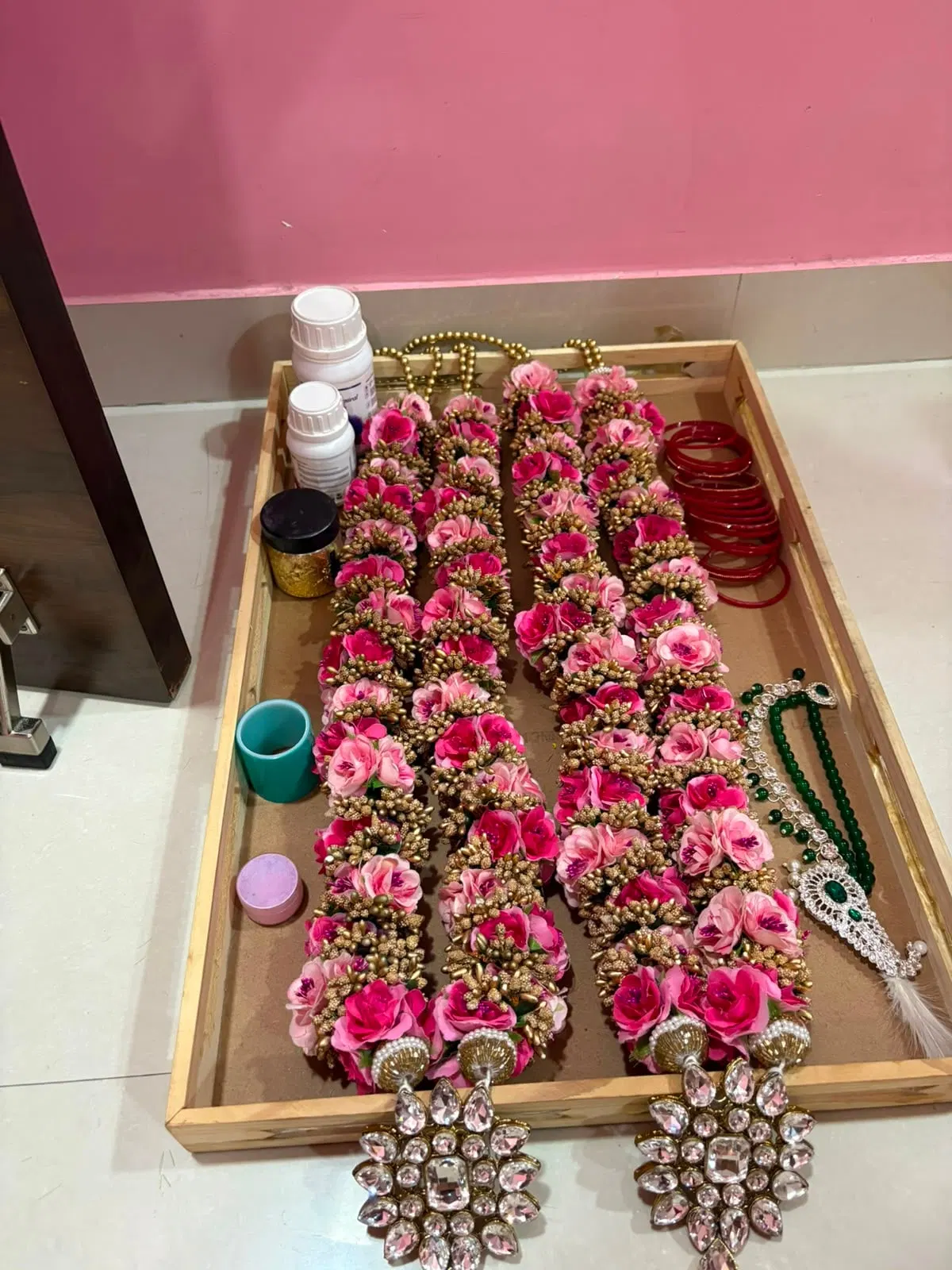 Two full pink-and-gold varmalas laid out in a wooden tray alongside red bangles, a green bead necklace and kundan pieces, with resin bottles and pigment pots beside them