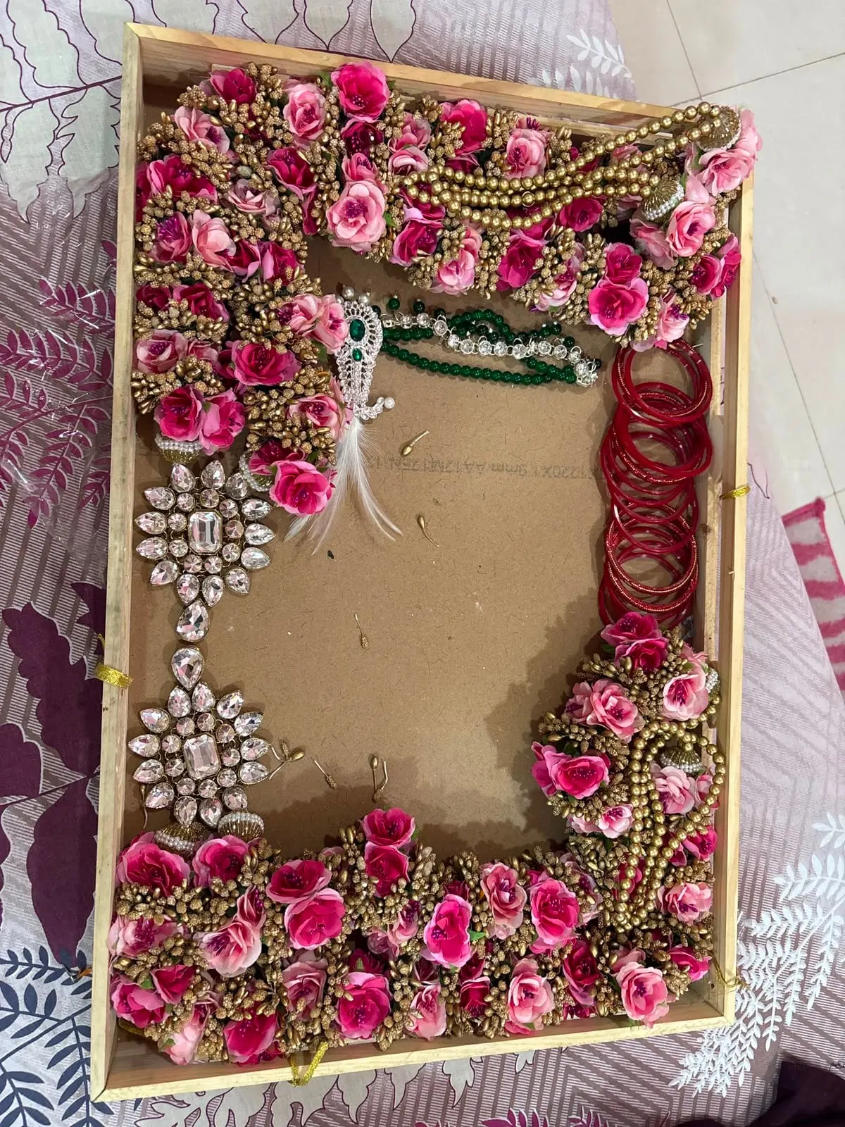 Overhead view of a varmala composition being laid out inside a wooden frame: roses forming a thick border with kundan brooches, a green bead necklace and red bangles placed inside, before the resin is poured