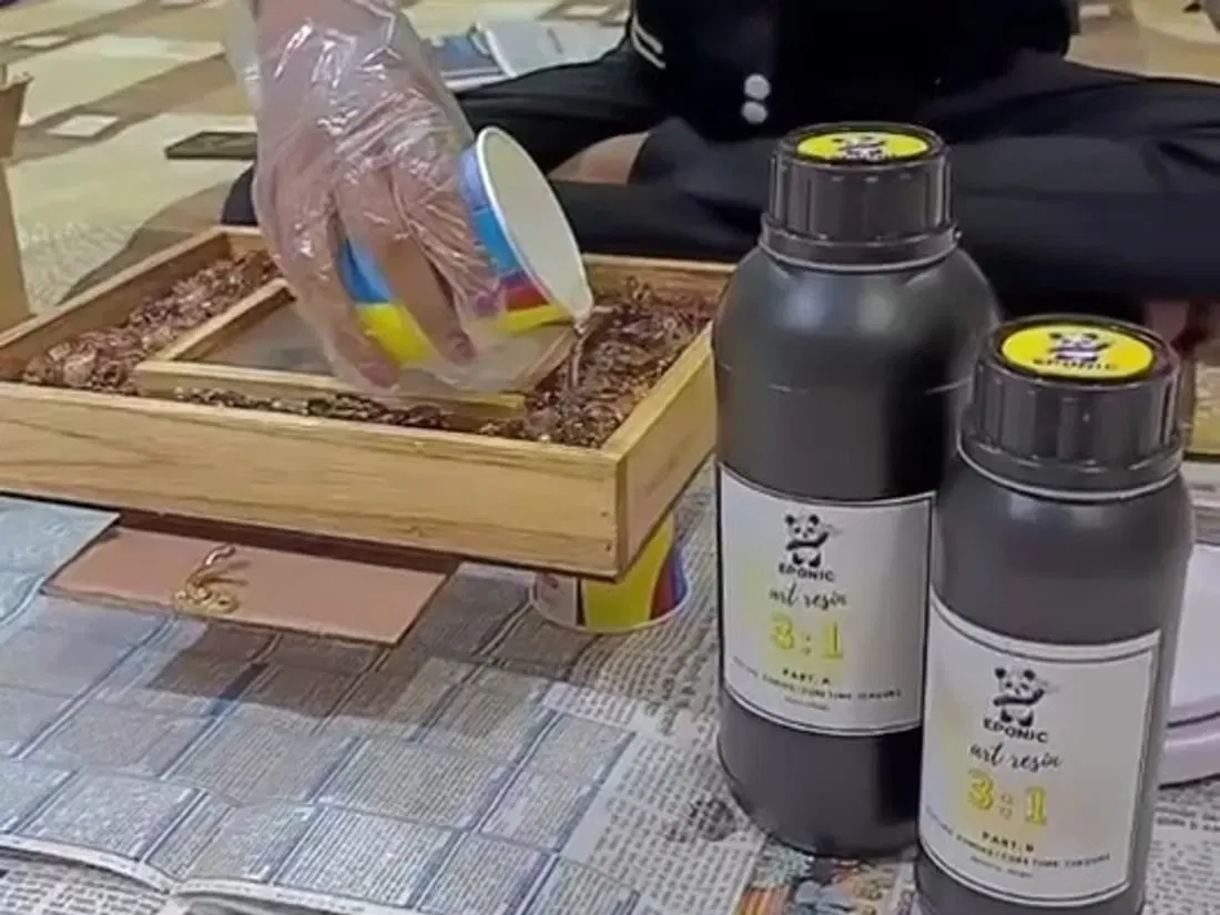 A gloved hand tipping mixed resin from a paper cup into a wooden frame of dried flowers, with two bottles of art resin standing on spread newspaper beside it